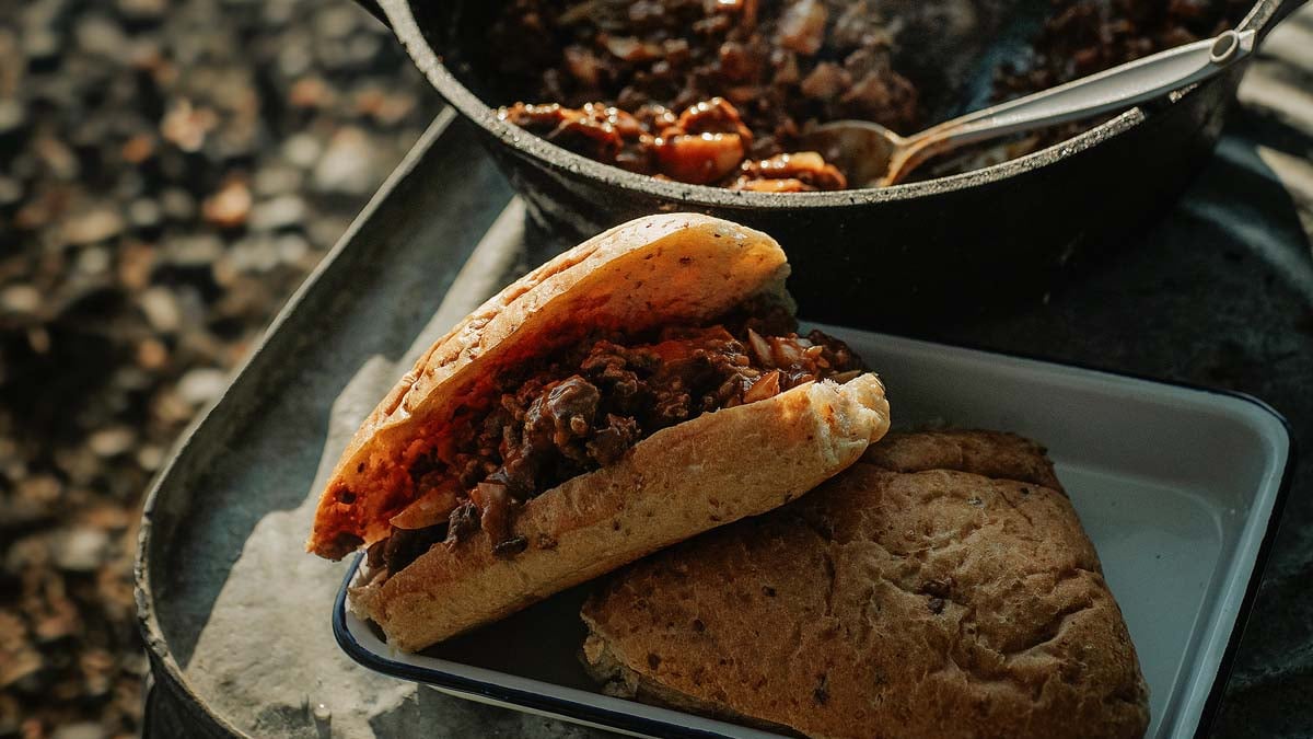 Campfire Sloppy Joes — Tangy, Make-Ahead Ground Beef for the Crowd 28 Close-up of a Campfire Sloppy Joe on a toasted bun with tangy beef filling, melted cheddar, and pickles, served on a rustic wooden board at a campsite
