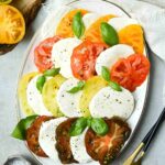 Beautiful Caprese Salad arranged on a white platter with sliced heirloom tomatoes, fresh mozzarella, basil leaves, and a drizzle of balsamic glaze and olive oil