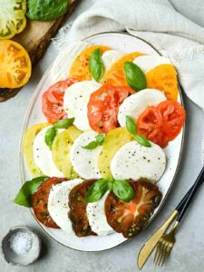 Beautiful Caprese Salad arranged on a white platter with sliced heirloom tomatoes, fresh mozzarella, basil leaves, and a drizzle of balsamic glaze and olive oil