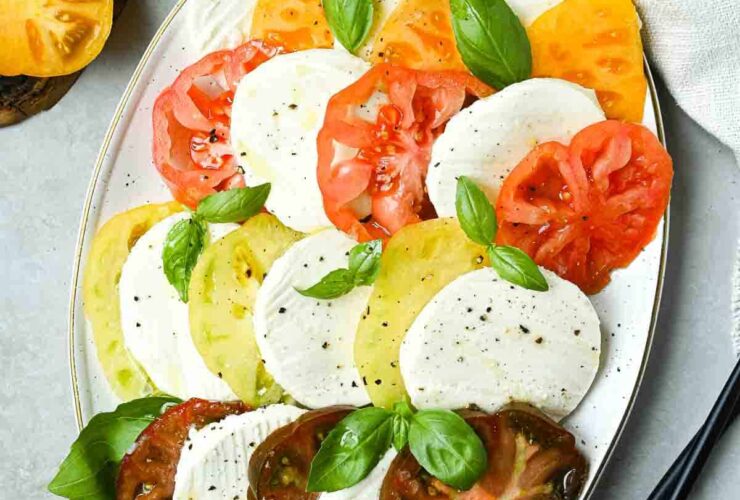 Beautiful Caprese Salad arranged on a white platter with sliced heirloom tomatoes, fresh mozzarella, basil leaves, and a drizzle of balsamic glaze and olive oil
