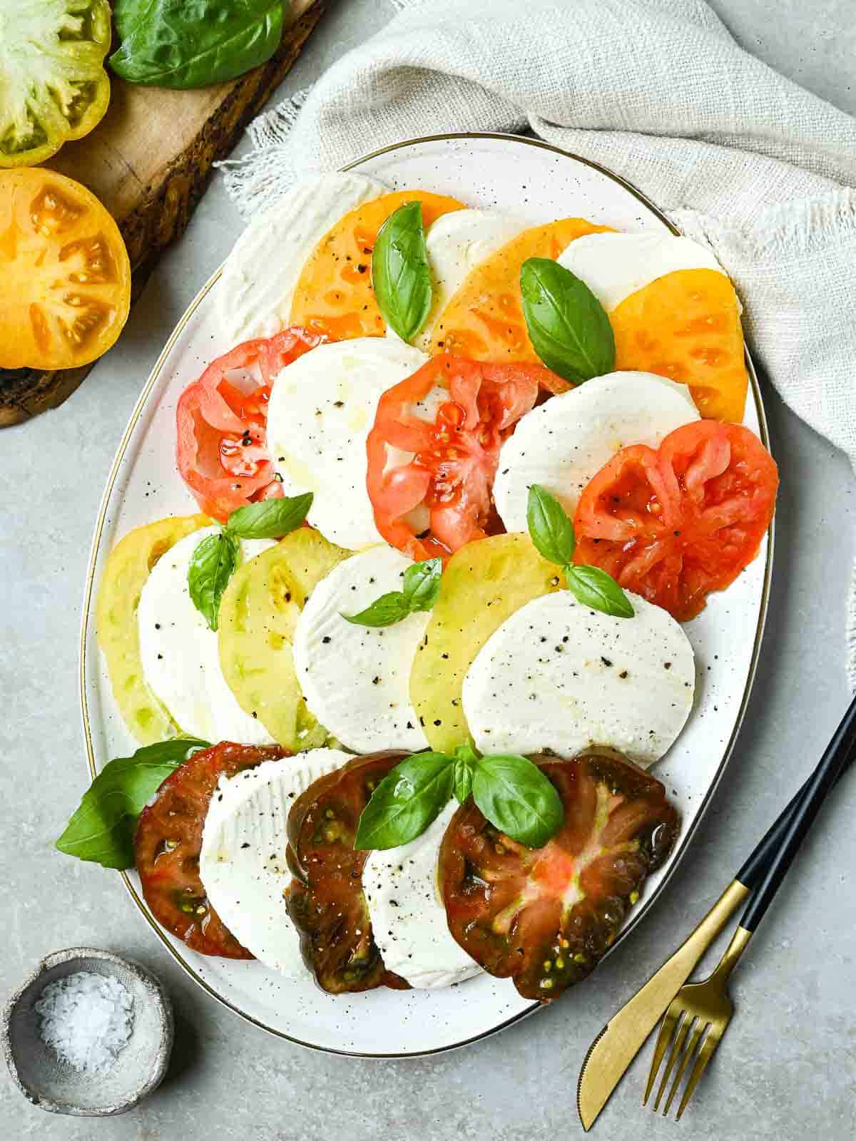 Beautiful Caprese Salad arranged on a white platter with sliced heirloom tomatoes, fresh mozzarella, basil leaves, and a drizzle of balsamic glaze and olive oil