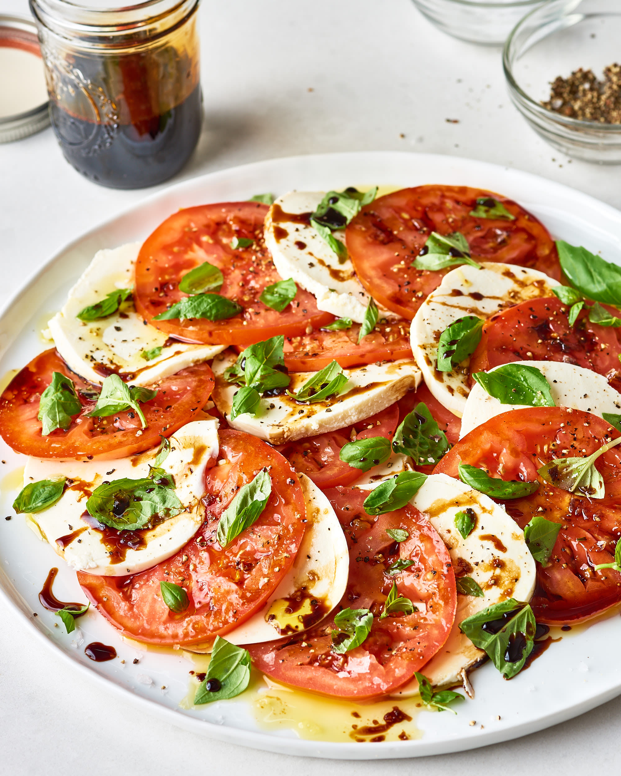 Overhead flat lay of Caprese Salad ingredients: heirloom tomatoes, fresh mozzarella ball, basil leaves, olive oil bottle, balsamic glaze, and flaky sea salt on a marble surface