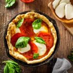 Cast iron skillet pizza with golden crispy crust, bubbling mozzarella cheese, and fresh basil leaves, photographed outdoors near a campfire