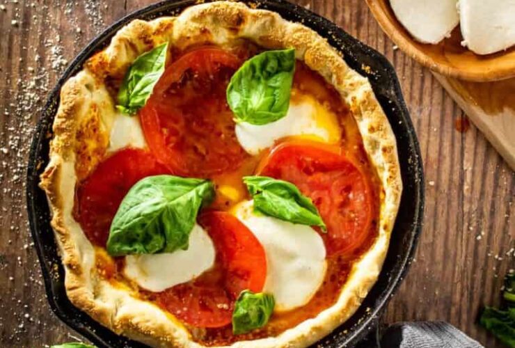 Cast iron skillet pizza with golden crispy crust, bubbling mozzarella cheese, and fresh basil leaves, photographed outdoors near a campfire