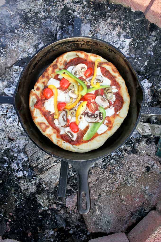 Step-by-step process of pressing pizza dough into a cast iron skillet outdoors, with toppings laid out on a camp table beside it