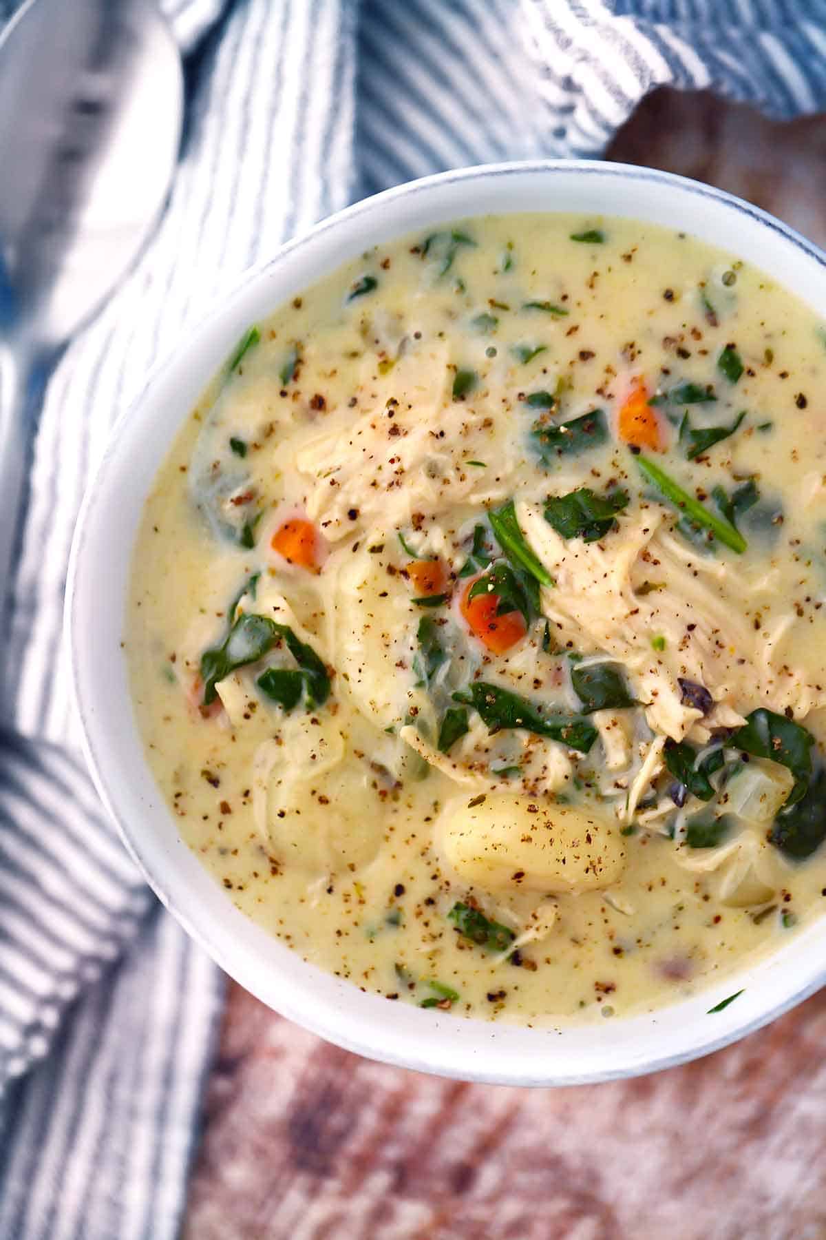 A rustic white bowl filled with creamy chicken gnocchi soup, pillowy gnocchi, shredded carrots, and spinach, garnished with cracked black pepper and fresh thyme, photographed from above on a warm linen surface