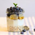 Coconut chia pudding parfait in a tall glass jar layered with mango, pineapple, toasted coconut flakes, and a drizzle of honey on a bright white marble surface with tropical greenery in the background