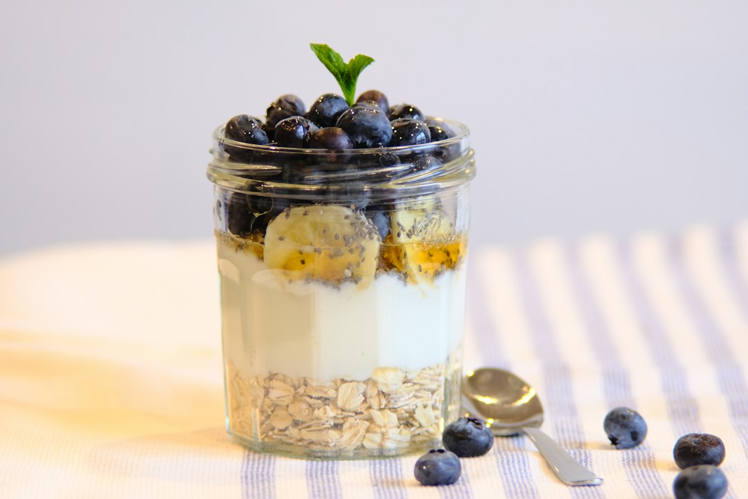 Coconut chia pudding parfait in a tall glass jar layered with mango, pineapple, toasted coconut flakes, and a drizzle of honey on a bright white marble surface with tropical greenery in the background