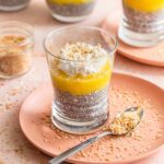 Coconut chia pudding parfait in a tall glass jar layered with mango, pineapple, toasted coconut flakes, and a drizzle of honey on a bright white marble surface with tropical greenery in the background