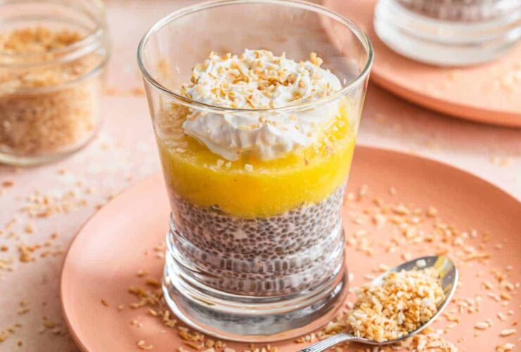 Coconut chia pudding parfait in a tall glass jar layered with mango, pineapple, toasted coconut flakes, and a drizzle of honey on a bright white marble surface with tropical greenery in the background