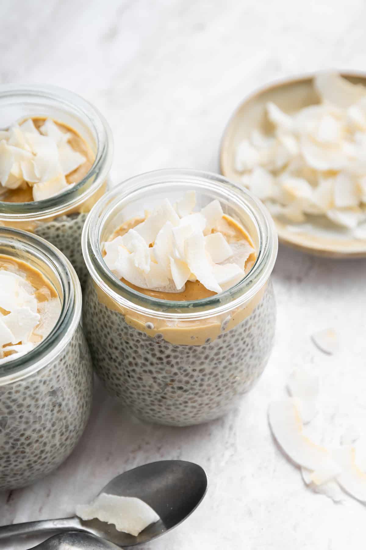 Close-up overhead shot of a coconut chia pudding parfait in a mason jar showing distinct layers of creamy white chia pudding, vibrant yellow mango cubes, pineapple chunks, and toasted golden coconut flakes with a small silver spoon resting beside it on a linen napkin