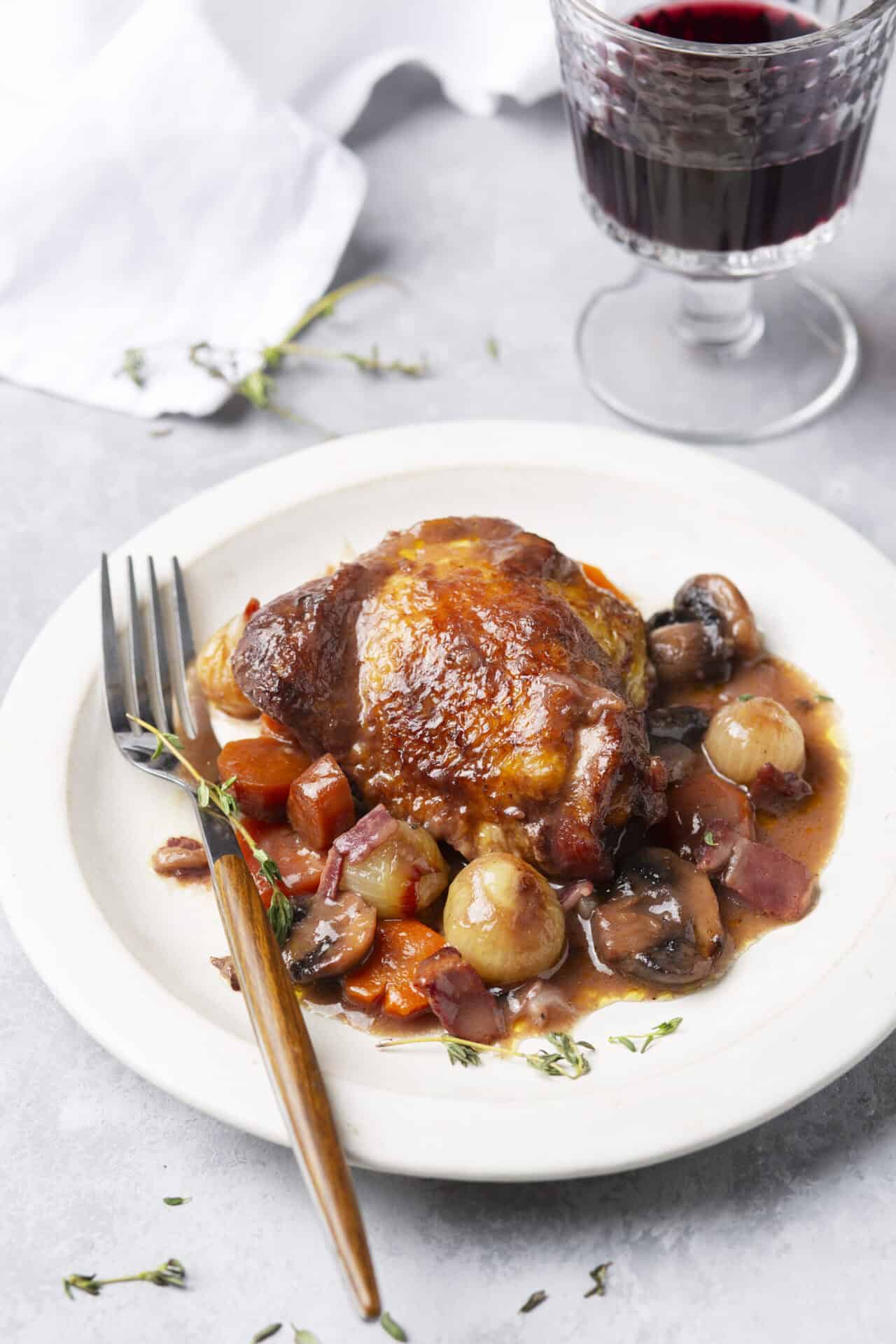 Close-up of a piece of Coq au Vin chicken thigh on a white plate with glossy red wine sauce, pearl onions, and fresh thyme sprig, fork resting alongside