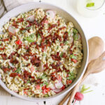 A large white serving bowl filled with creamy macaroni salad garnished with paprika and fresh parsley, photographed overhead on a rustic wooden picnic table with a checkered napkin