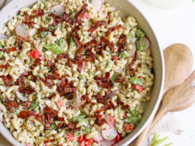 A large white serving bowl filled with creamy macaroni salad garnished with paprika and fresh parsley, photographed overhead on a rustic wooden picnic table with a checkered napkin