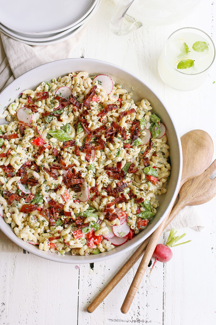 A large white serving bowl filled with creamy macaroni salad garnished with paprika and fresh parsley, photographed overhead on a rustic wooden picnic table with a checkered napkin