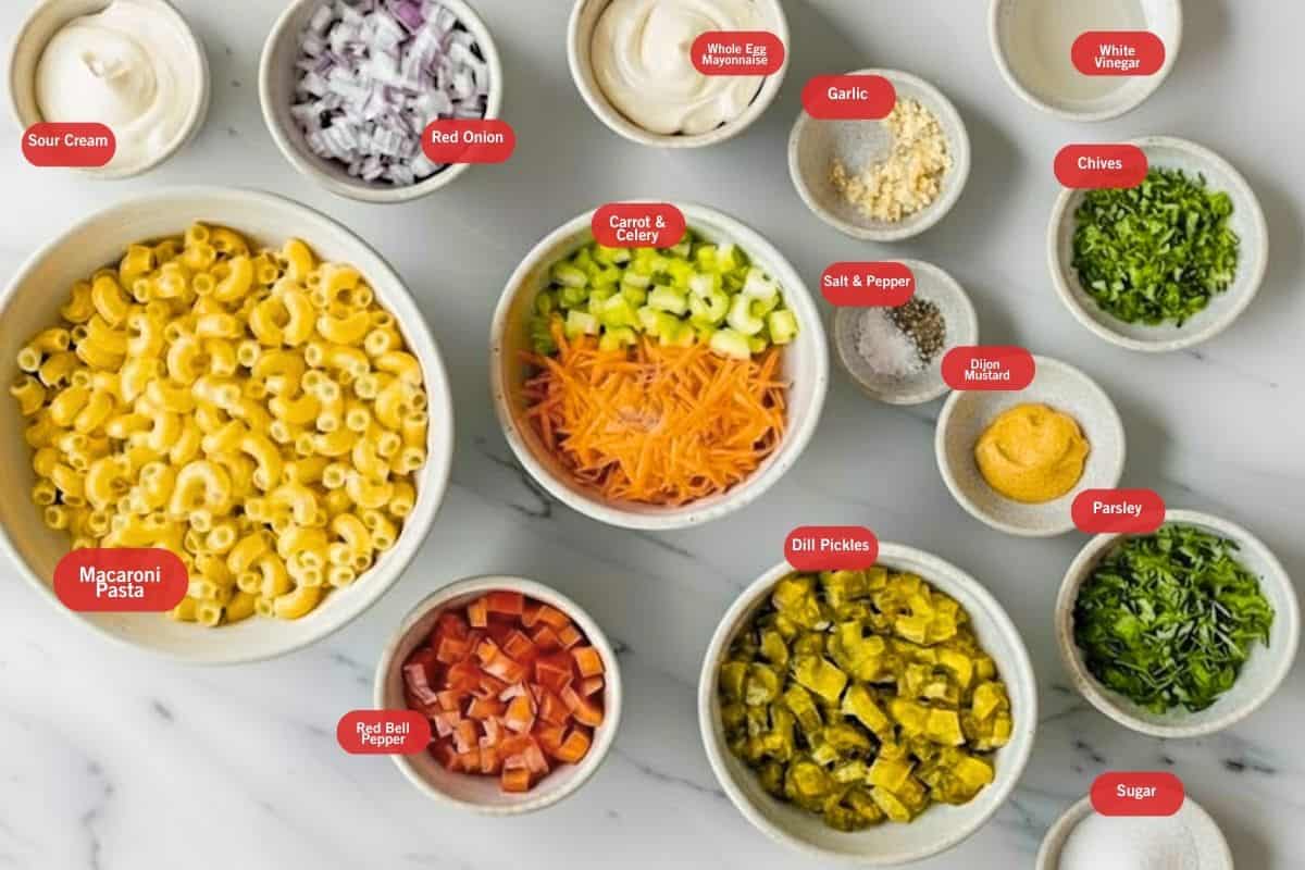 A flat lay of creamy macaroni salad ingredients — elbow pasta, celery, red bell pepper, red onion, mayonnaise, Dijon mustard, and apple cider vinegar arranged on a clean white surface