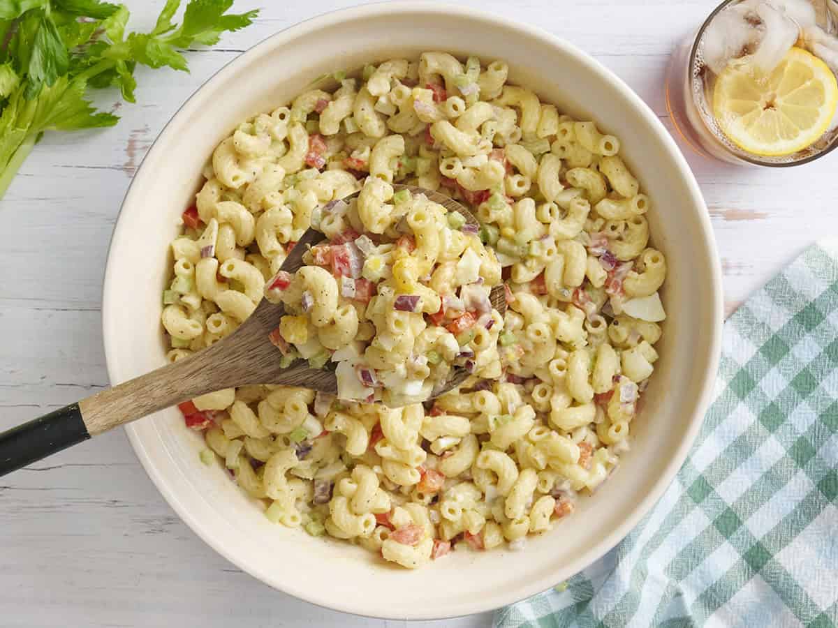A close-up shot of a spoonful of creamy macaroni salad being lifted from the bowl, showing the glossy mayo dressing, colorful vegetable pieces, and perfectly coated pasta
