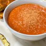 A white ceramic bowl filled with deep red creamy tomato basil soup, garnished with a swirl of cream, fresh basil leaves, and a crack of black pepper, with a golden grilled cheese sandwich resting on the side of the bowl