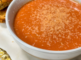 A white ceramic bowl filled with deep red creamy tomato basil soup, garnished with a swirl of cream, fresh basil leaves, and a crack of black pepper, with a golden grilled cheese sandwich resting on the side of the bowl