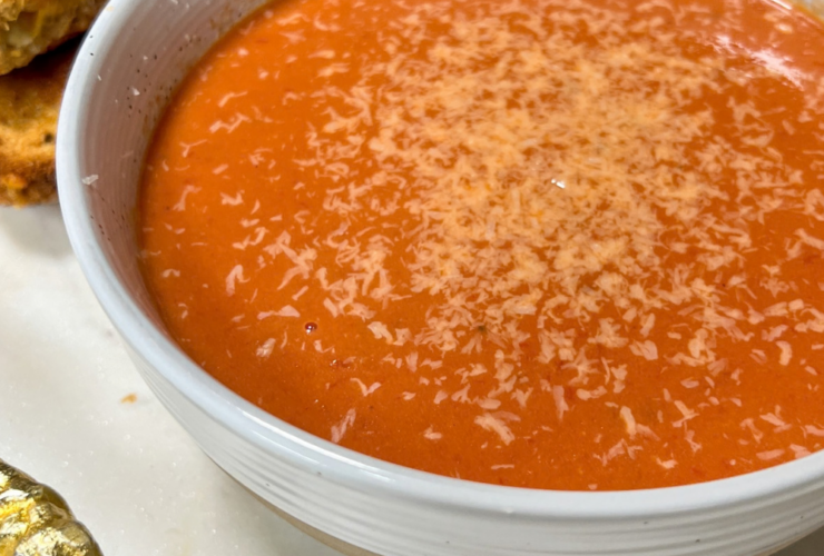 A white ceramic bowl filled with deep red creamy tomato basil soup, garnished with a swirl of cream, fresh basil leaves, and a crack of black pepper, with a golden grilled cheese sandwich resting on the side of the bowl