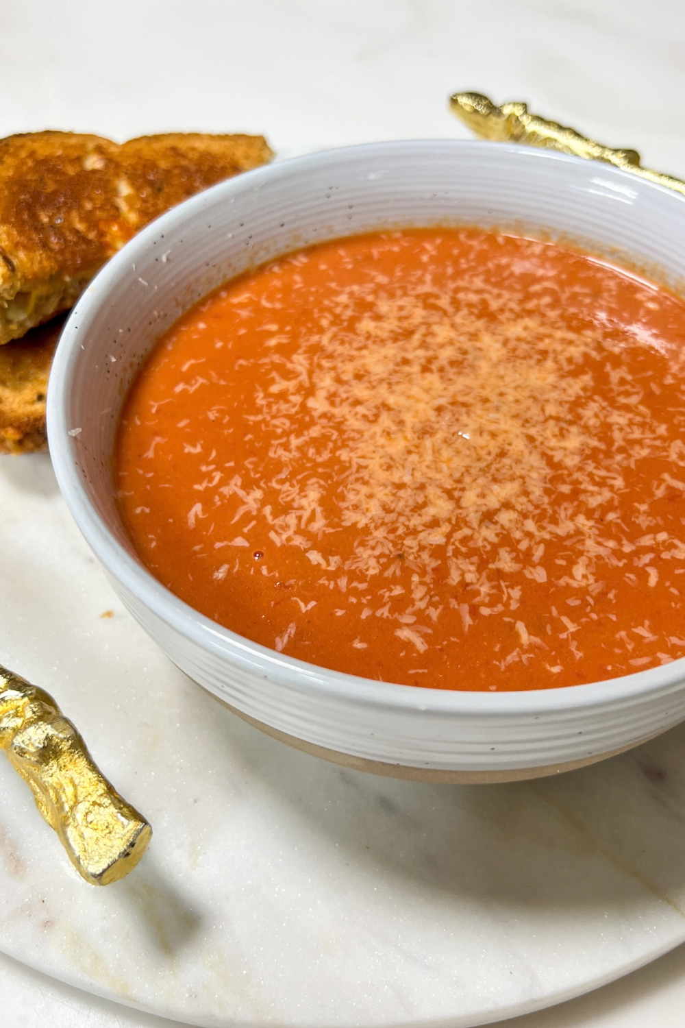 Creamy Tomato Basil Soup — Roasted Tomato Soup Built for Grilled Cheese Dunking 26 A white ceramic bowl filled with deep red creamy tomato basil soup, garnished with a swirl of cream, fresh basil leaves, and a crack of black pepper, with a golden grilled cheese sandwich resting on the side of the bowl