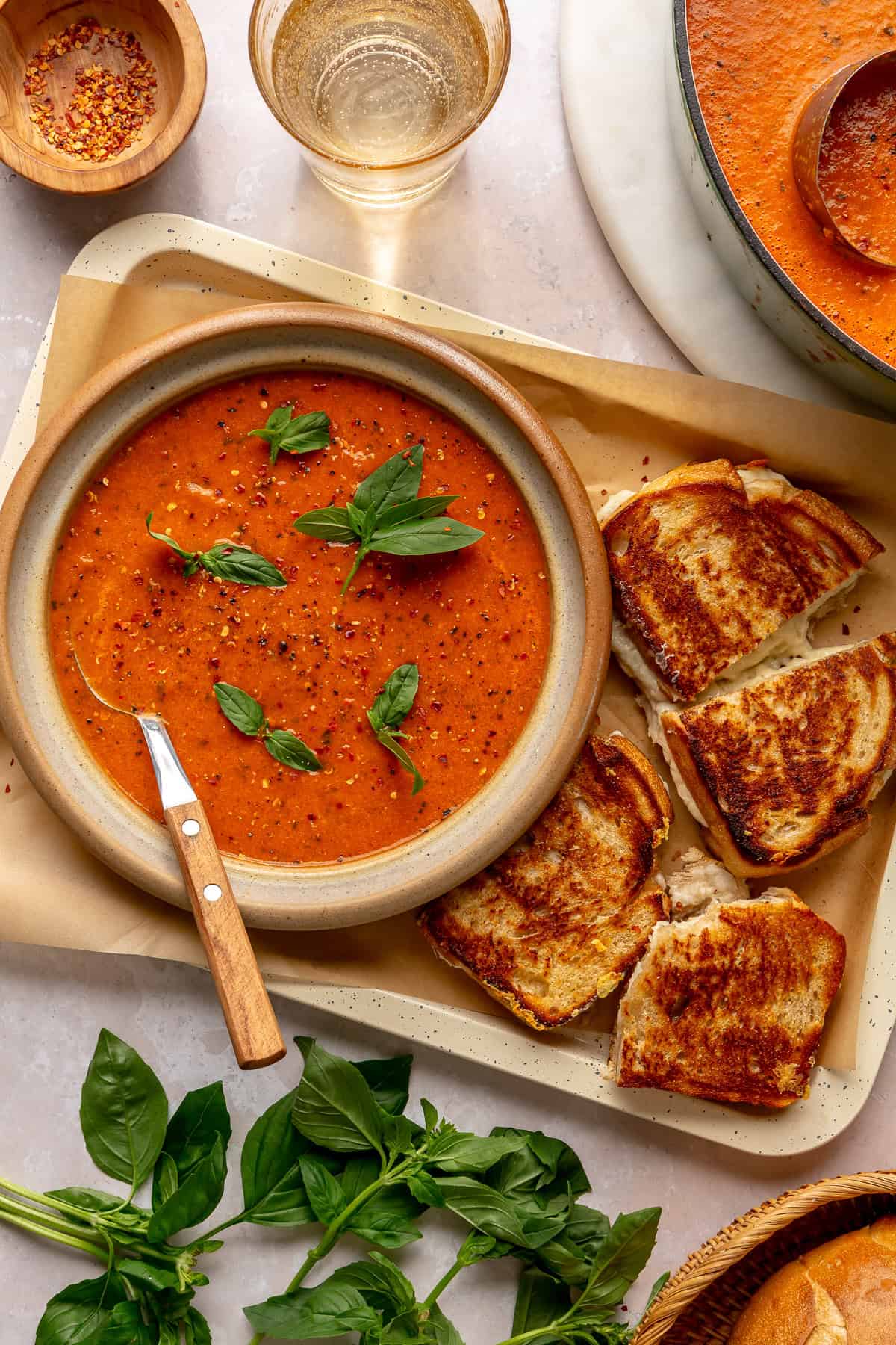 Creamy Tomato Basil Soup — Roasted Tomato Soup Built for Grilled Cheese Dunking 28 A close-up overhead shot of a bowl of creamy tomato basil soup with a golden grilled cheese sandwich cut diagonally, one half being dunked into the soup, with fresh basil scattered around the bowl on a rustic wooden surface