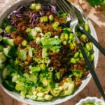Crispy rice salad served in a wide shallow bowl with golden pan-fried rice, sliced cucumbers, edamame, shredded purple cabbage, avocado, and drizzled with sesame dressing, garnished with sesame seeds and fresh cilantro