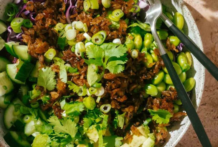 Crispy rice salad served in a wide shallow bowl with golden pan-fried rice, sliced cucumbers, edamame, shredded purple cabbage, avocado, and drizzled with sesame dressing, garnished with sesame seeds and fresh cilantro