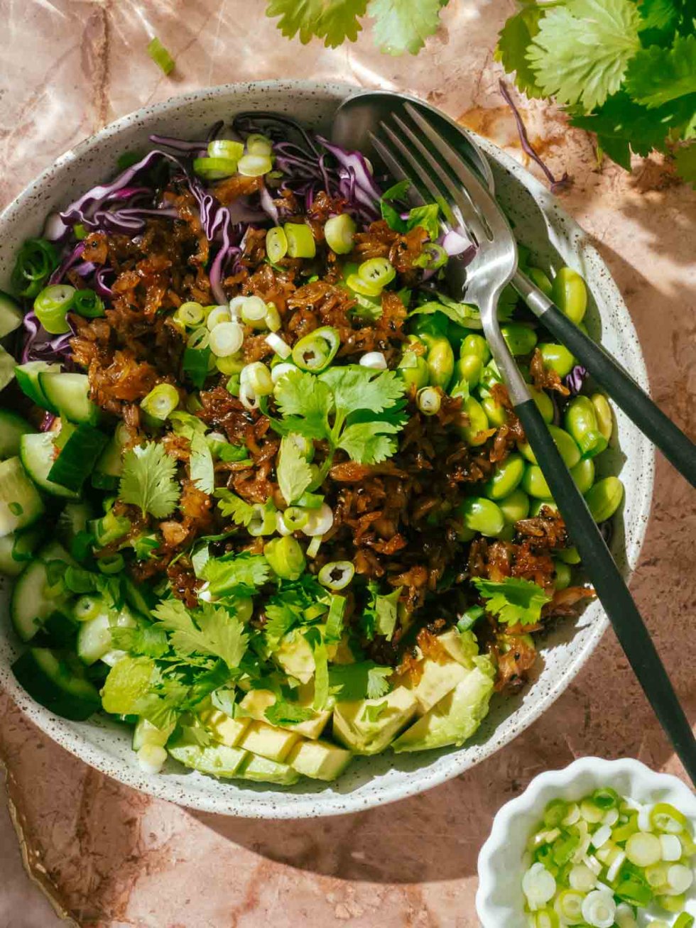 Close-up of a serving of crispy rice salad in a ceramic bowl, showing golden rice clusters glistening with sesame dressing, surrounded by vibrant green edamame, cucumber coins, and creamy avocado slices topped with sesame seeds and lime wedge