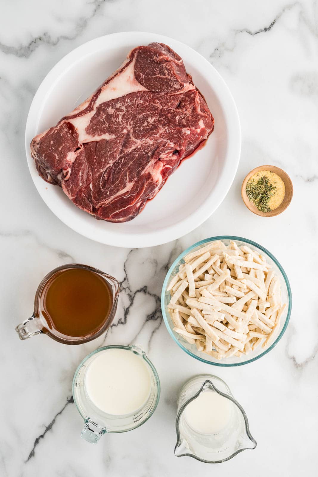 Overhead flat-lay of crockpot beef and noodles ingredients including chuck roast, egg noodles, beef broth, garlic, onion, and Worcestershire sauce arranged on a wooden board