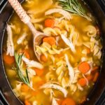 A steaming bowl of Crockpot Chicken Noodle Soup with golden broth, tender shredded chicken, thick egg noodles, and sliced carrots, garnished with fresh parsley, photographed overhead on a rustic wooden table