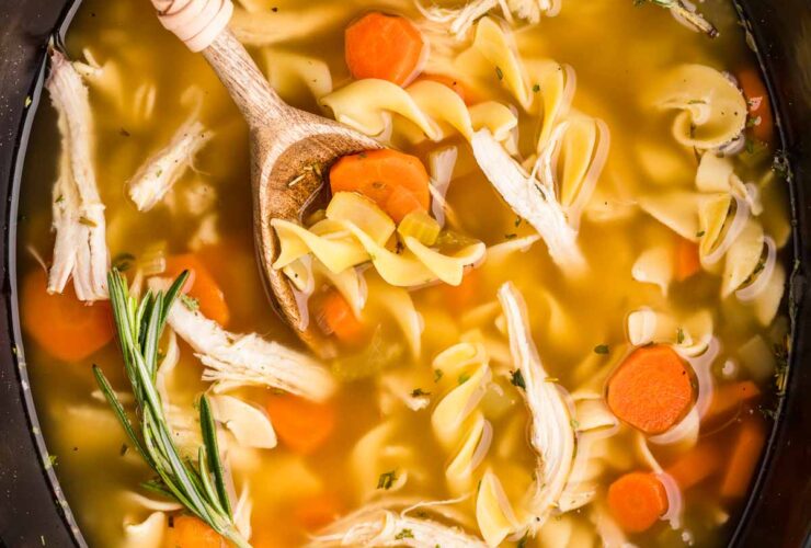 A steaming bowl of Crockpot Chicken Noodle Soup with golden broth, tender shredded chicken, thick egg noodles, and sliced carrots, garnished with fresh parsley, photographed overhead on a rustic wooden table