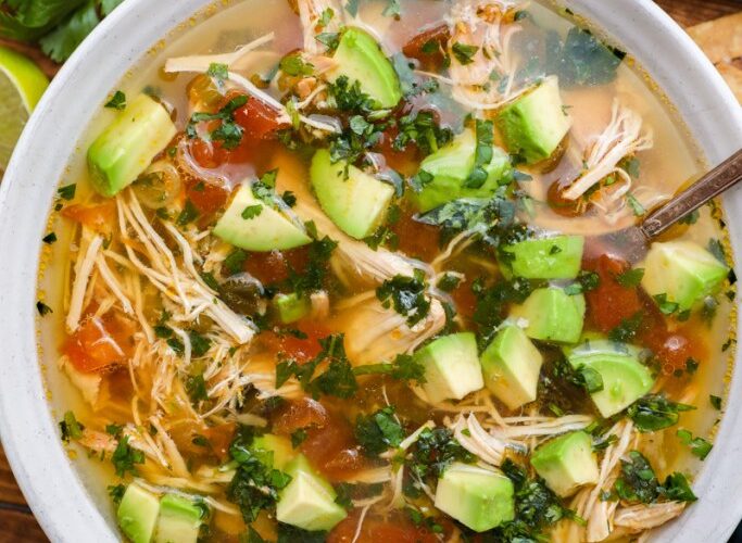 Crockpot Chicken Tortilla Soup served in a rustic bowl topped with tortilla strips, avocado, shredded cheese, cilantro, and a lime wedge on a warm wooden table