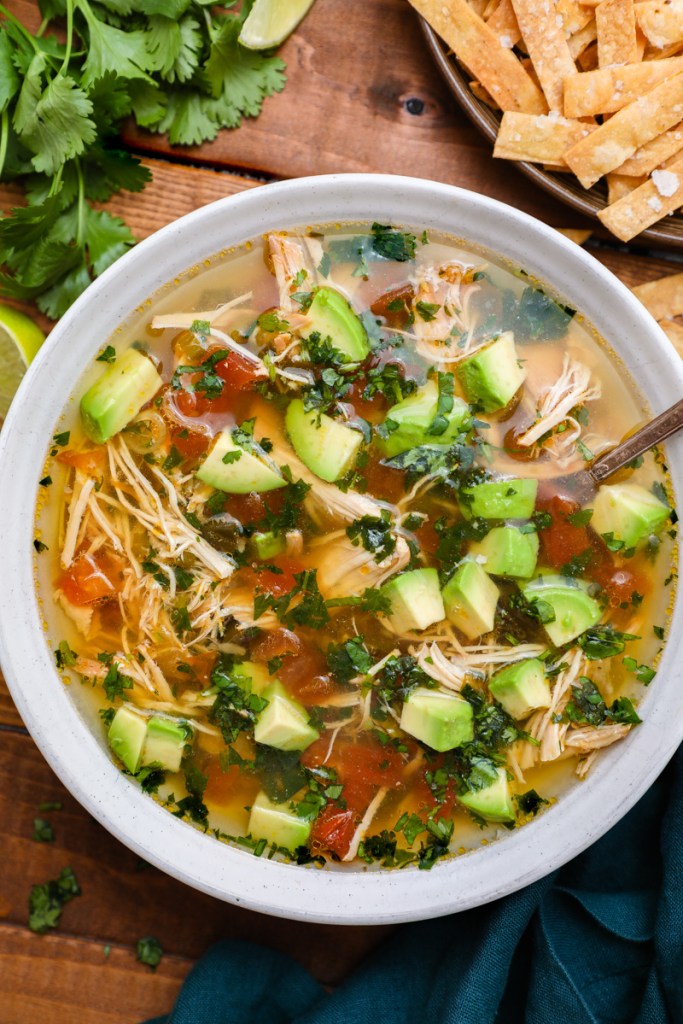 Crockpot Chicken Tortilla Soup served in a rustic bowl topped with tortilla strips, avocado, shredded cheese, cilantro, and a lime wedge on a warm wooden table