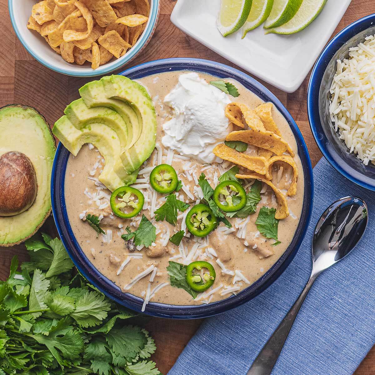 Crockpot White Chicken Chili — Creamy, Cozy, and Packed with Protein 28 A close-up overhead shot of a white ceramic bowl filled with Crockpot White Chicken Chili, garnished with creamy avocado slices, bright green cilantro leaves, a wedge of lime, and a swirl of sour cream, with tortilla chips resting on the side of the bowl