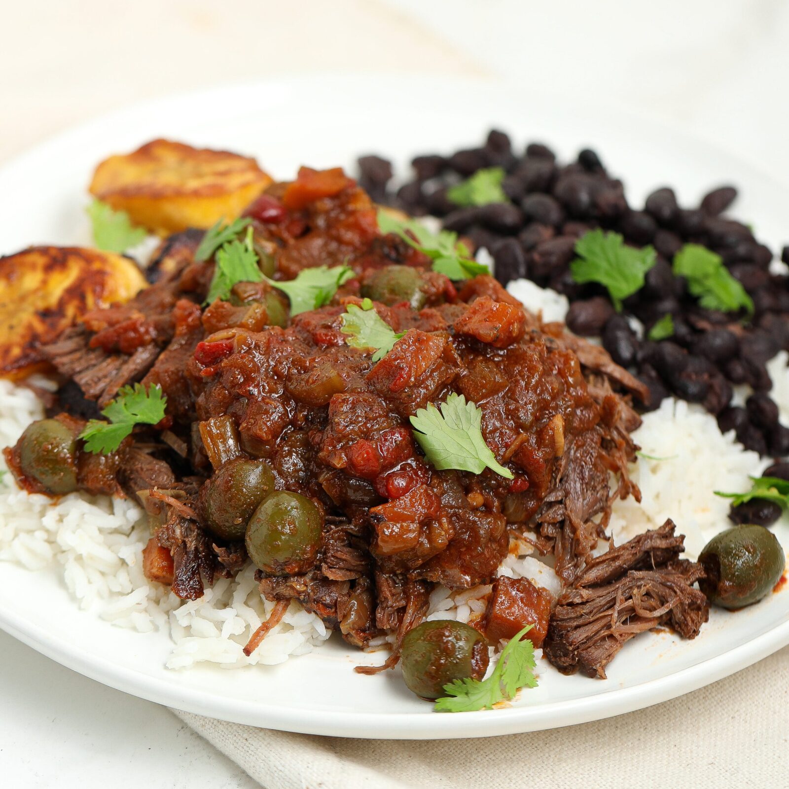 Cuban Ropa Vieja — Tender Shredded Beef in Rich Tomato Sofrito Sauce 25 Cuban Ropa Vieja served over white rice in a wide bowl, topped with fresh cilantro and sliced olives, photographed on a rustic wooden table with warm lighting