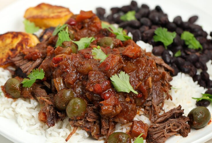 Cuban Ropa Vieja — Tender Shredded Beef in Rich Tomato Sofrito Sauce 27 Cuban Ropa Vieja served over white rice in a wide bowl, topped with fresh cilantro and sliced olives, photographed on a rustic wooden table with warm lighting