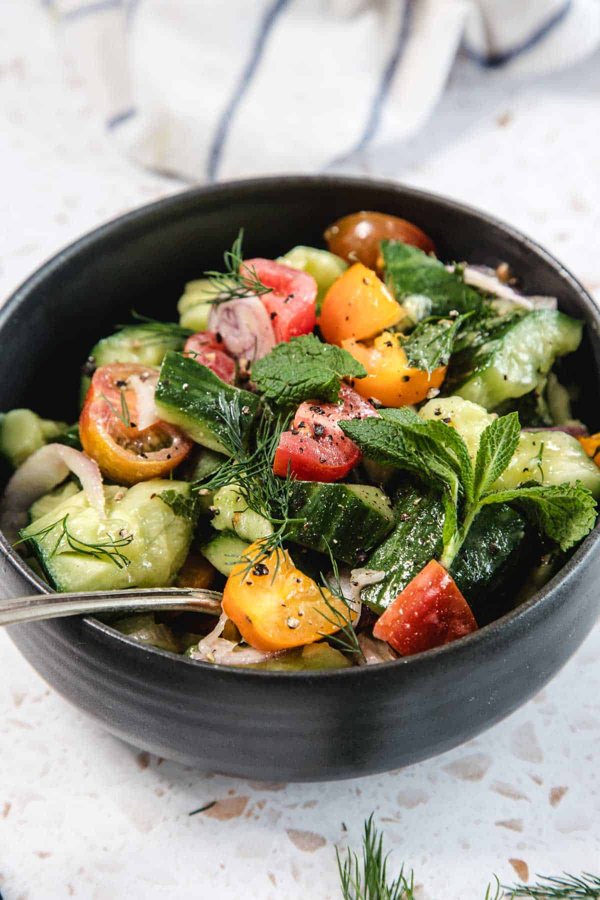 Sliced cucumbers, halved tomatoes, thinly sliced red onion, and fresh dill arranged on a wooden cutting board beside a small bowl of vinegar dressing