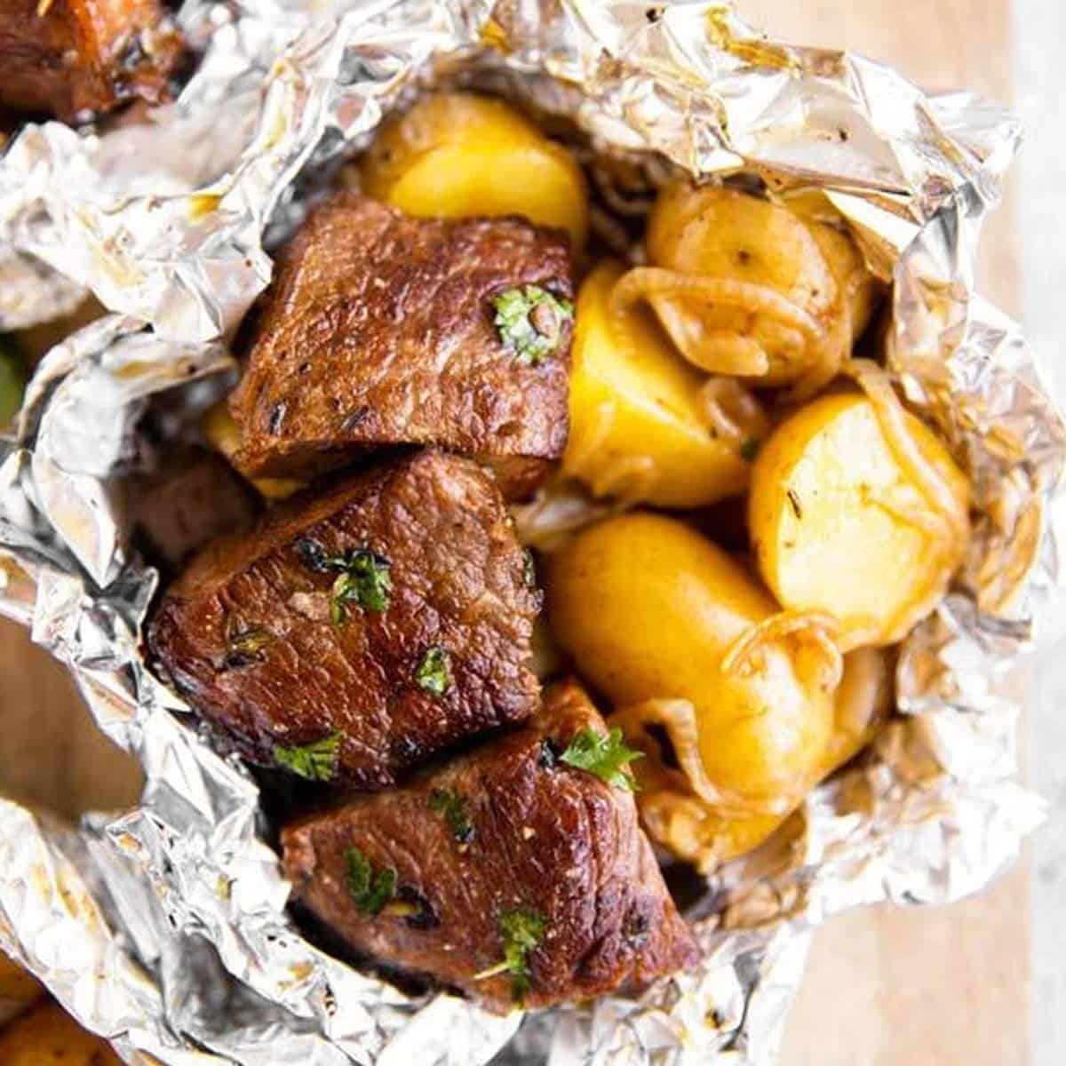 Close-up of Foil Packet Steak and Potato Hash opened at the table, steam rising, with fork-tender steak pieces glistening alongside golden potato halves and crispy bacon, topped with a sprinkle of fresh parsley