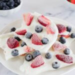 Frozen Greek yogurt berry bark broken into pieces on a white marble surface, topped with fresh strawberries, blueberries, and raspberries, drizzled with honey, bright natural lighting