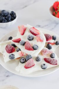 Frozen Greek Yogurt Berry Bark — The Healthy Dessert You'll Make All Summer 13 Frozen Greek yogurt berry bark broken into pieces on a white marble surface, topped with fresh strawberries, blueberries, and raspberries, drizzled with honey, bright natural lighting