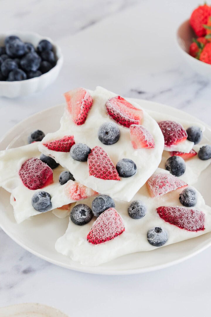 Frozen Greek yogurt berry bark broken into pieces on a white marble surface, topped with fresh strawberries, blueberries, and raspberries, drizzled with honey, bright natural lighting