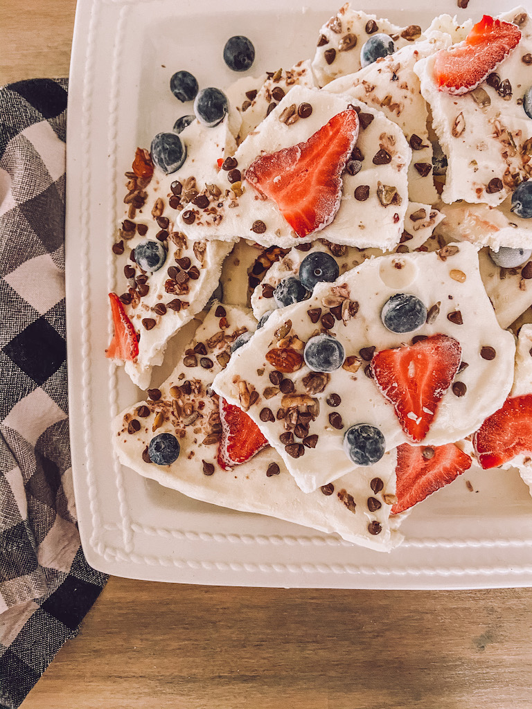 Overhead flat lay of ingredients for frozen Greek yogurt bark — bowl of thick Greek yogurt, colorful mixed berries, honey, granola, and dark chocolate chips arranged on a light wooden surface
