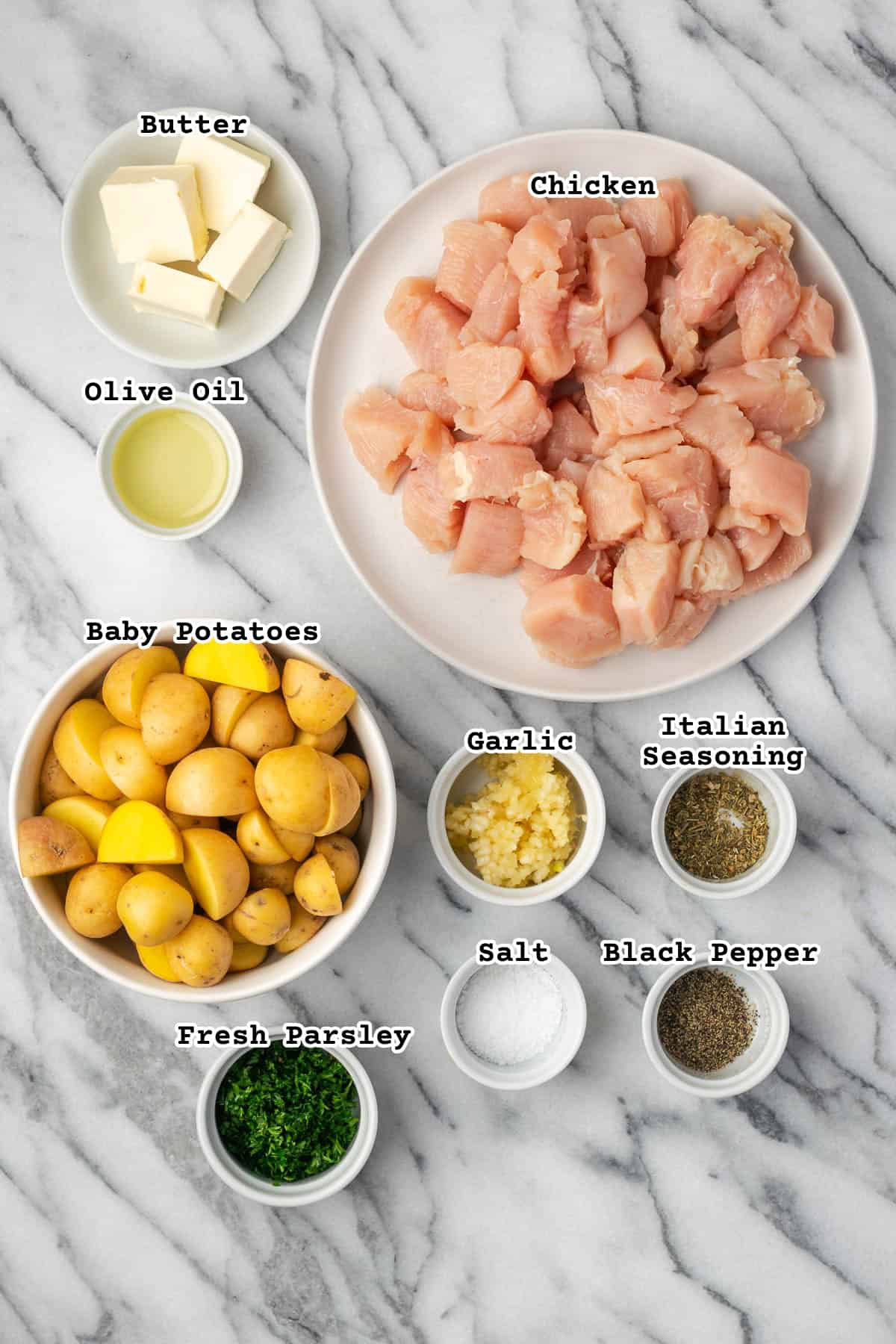 Overhead flat lay of raw chicken bites, halved baby potatoes, minced garlic, butter, smoked paprika, and fresh parsley arranged on a white marble surface