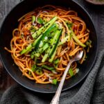 Gochujang noodles in a dark bowl garnished with sesame seeds, sliced scallions, and a soft-boiled egg, photographed overhead on a slate surface