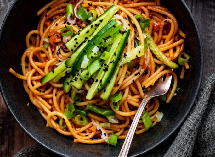 Gochujang noodles in a dark bowl garnished with sesame seeds, sliced scallions, and a soft-boiled egg, photographed overhead on a slate surface