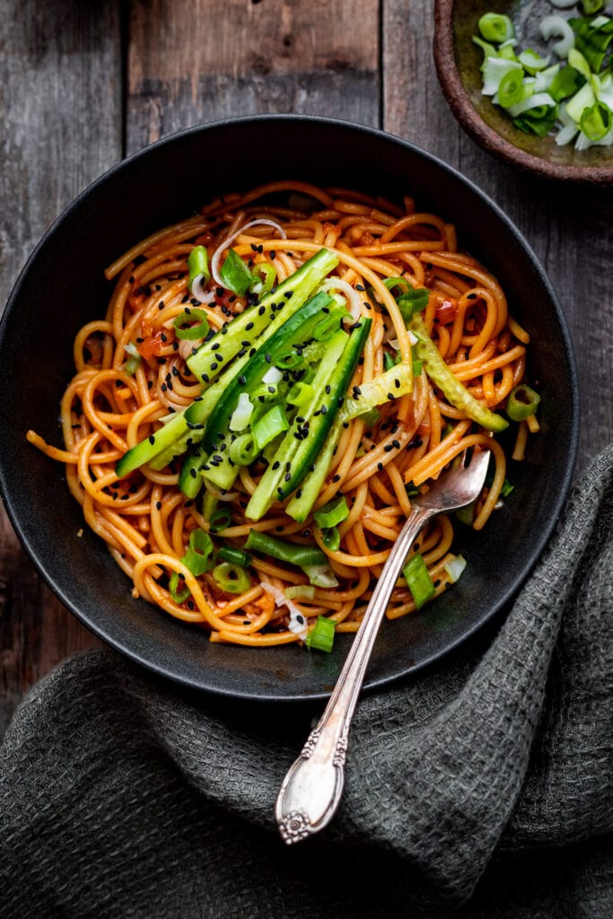 Gochujang noodles in a dark bowl garnished with sesame seeds, sliced scallions, and a soft-boiled egg, photographed overhead on a slate surface