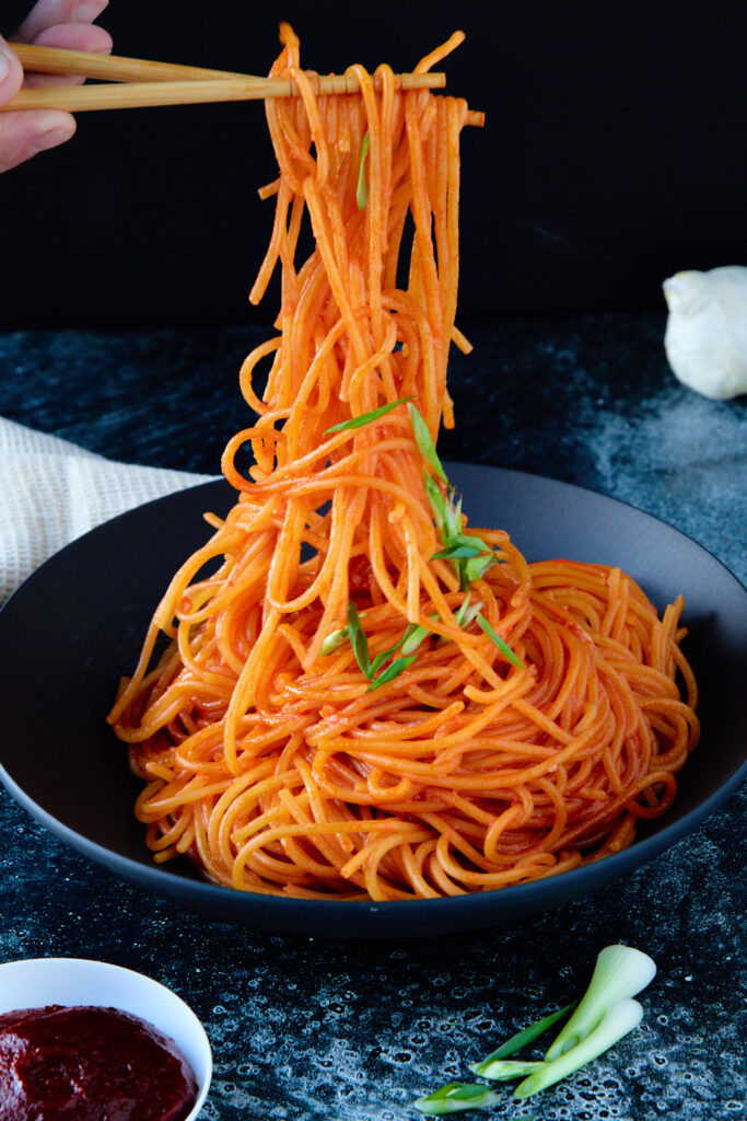 Close-up of gochujang noodles being lifted with chopsticks, showing the glossy sauce coating and sesame seeds, with a soft-boiled egg in the background