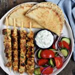 Greek Chicken Souvlaki served on a platter with warm pita, tzatziki, sliced tomatoes, red onion, and fresh parsley garnish