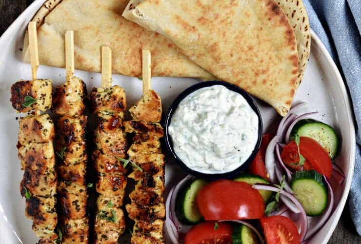Greek Chicken Souvlaki served on a platter with warm pita, tzatziki, sliced tomatoes, red onion, and fresh parsley garnish
